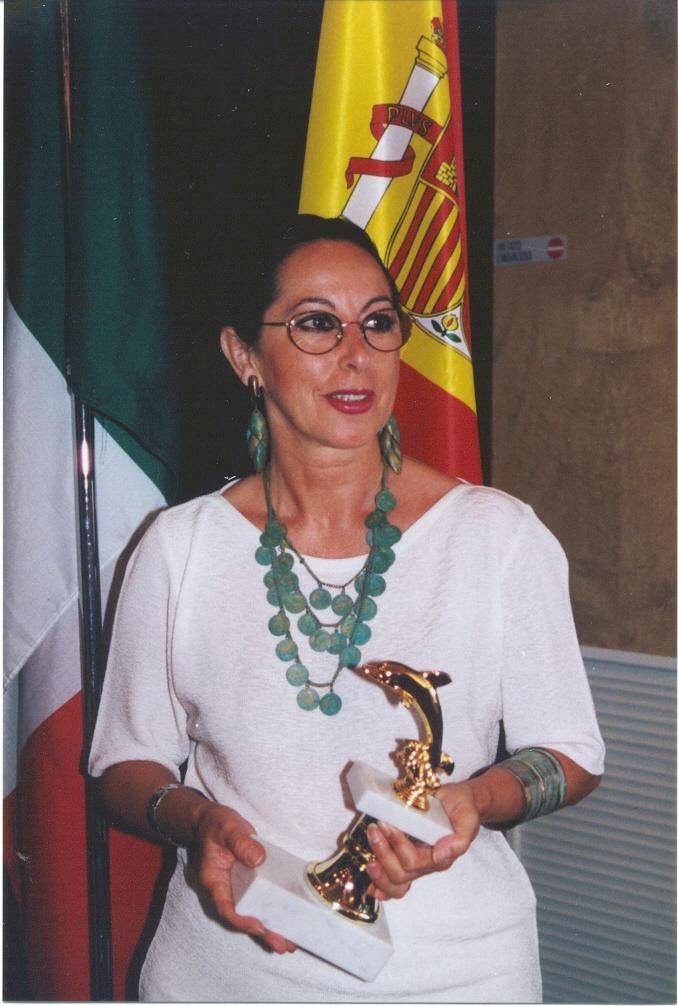 Mujer sosteniendo un trofeo, con gafas y joyas verdes, de pie entre banderas de Italia y España.