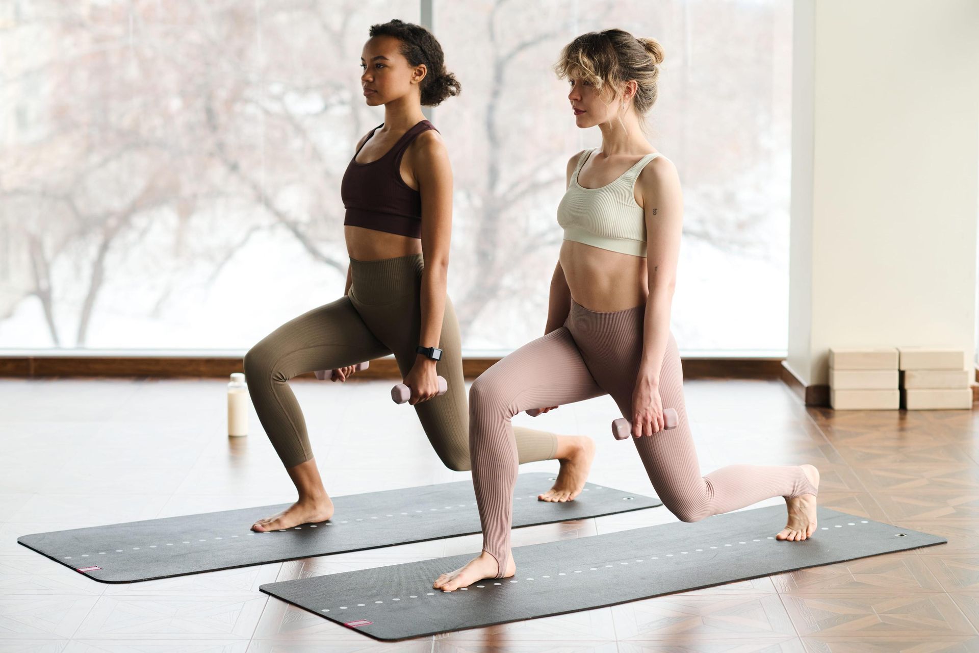 En un estudio luminoso, dos personas realizan zancadas sosteniendo pequeñas mancuernas sobre esterillas de yoga.