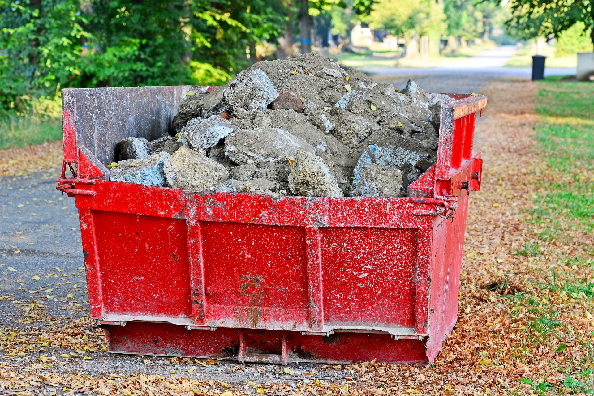 Une grande benne à ordures en métal rouge vif, remplie de terre, de pierres et de débris, trônait sur un chemin jonché de feuilles mortes d'automne.