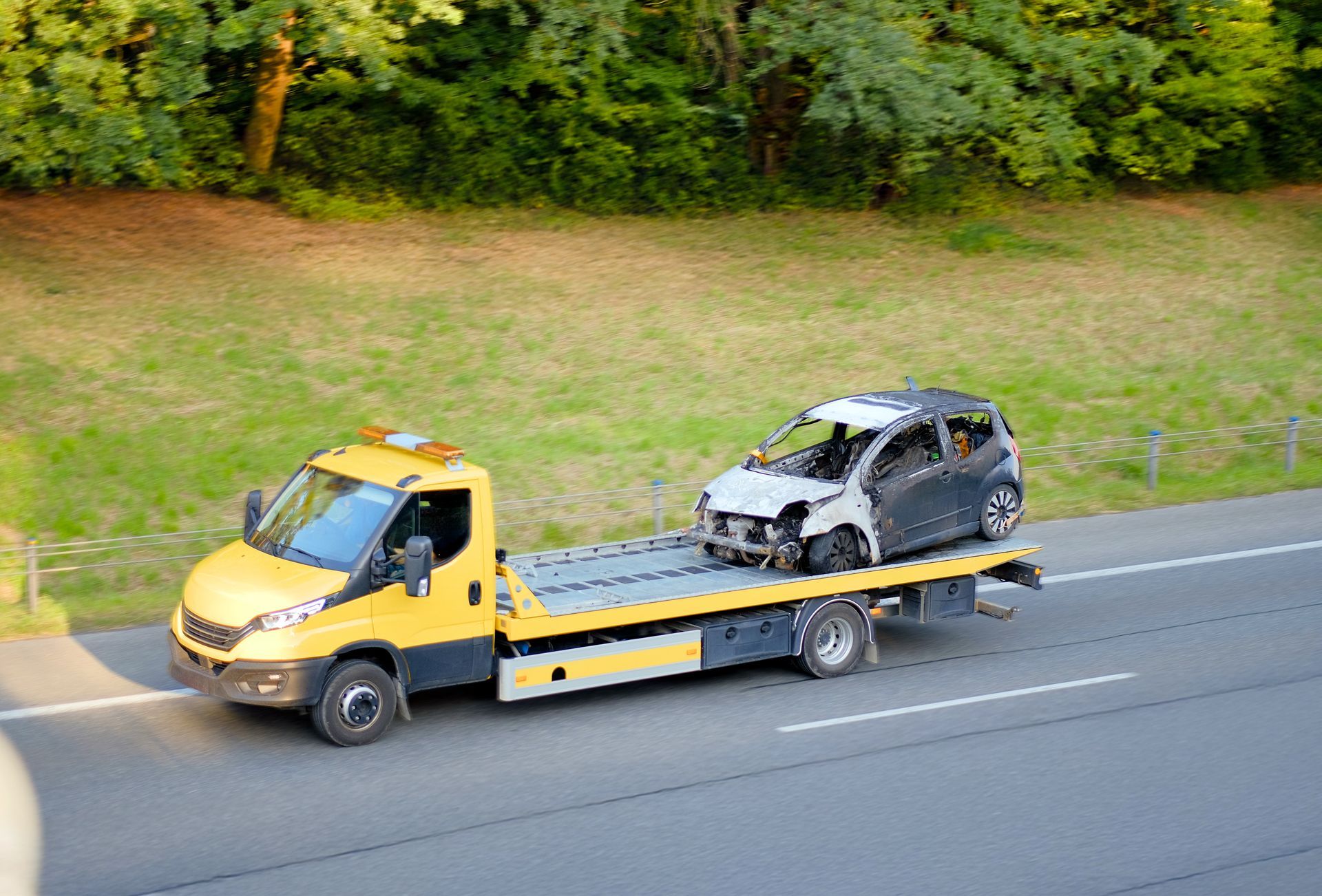 Une dépanneuse jaune à plateau transporte une petite voiture calcinée et calcinée le long d'une autoroute, à côté d'un talus herbeux.