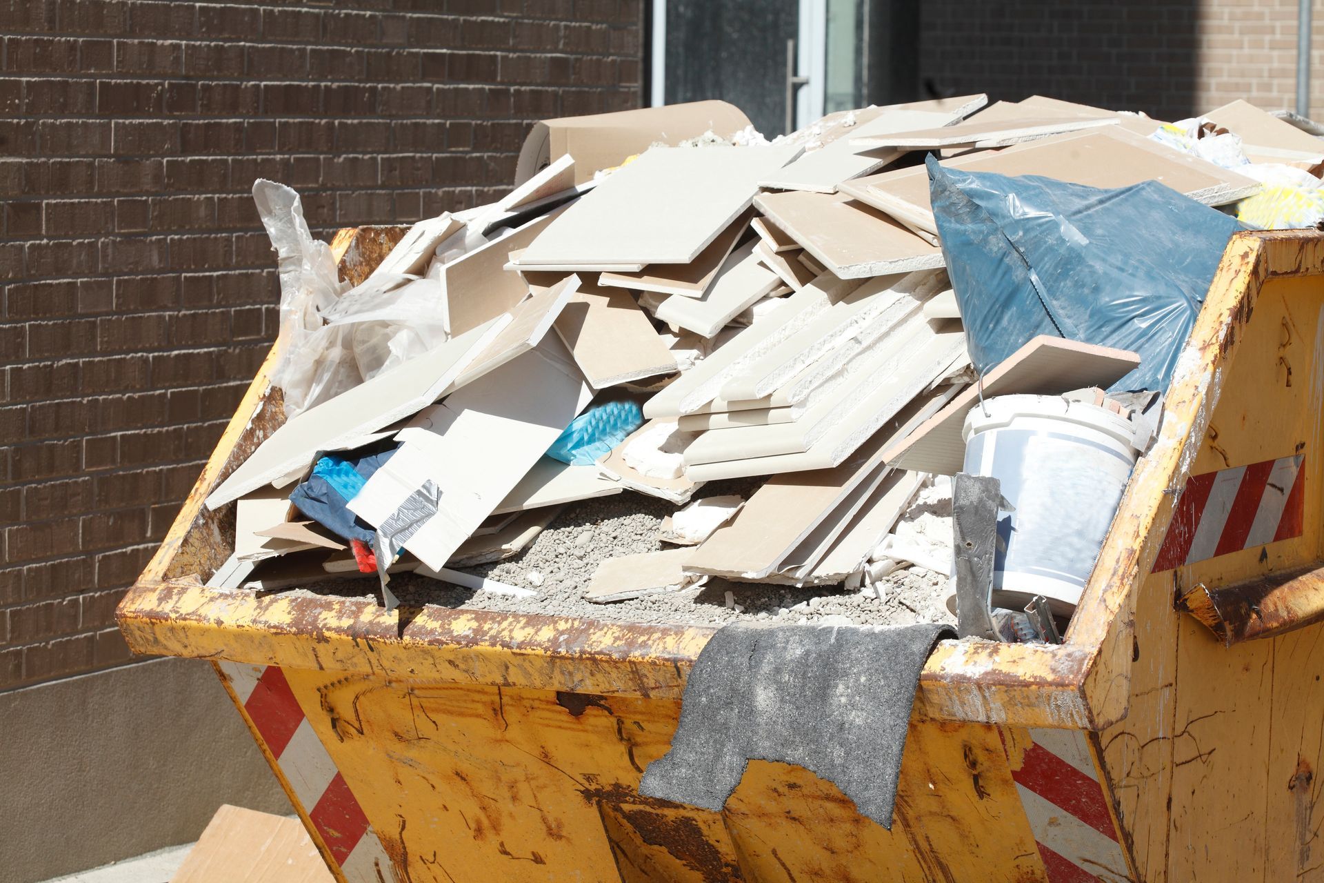 Une benne à ordures jaune remplie de débris, notamment des chutes de plaques de plâtre, un sac bleu et un seau blanc.