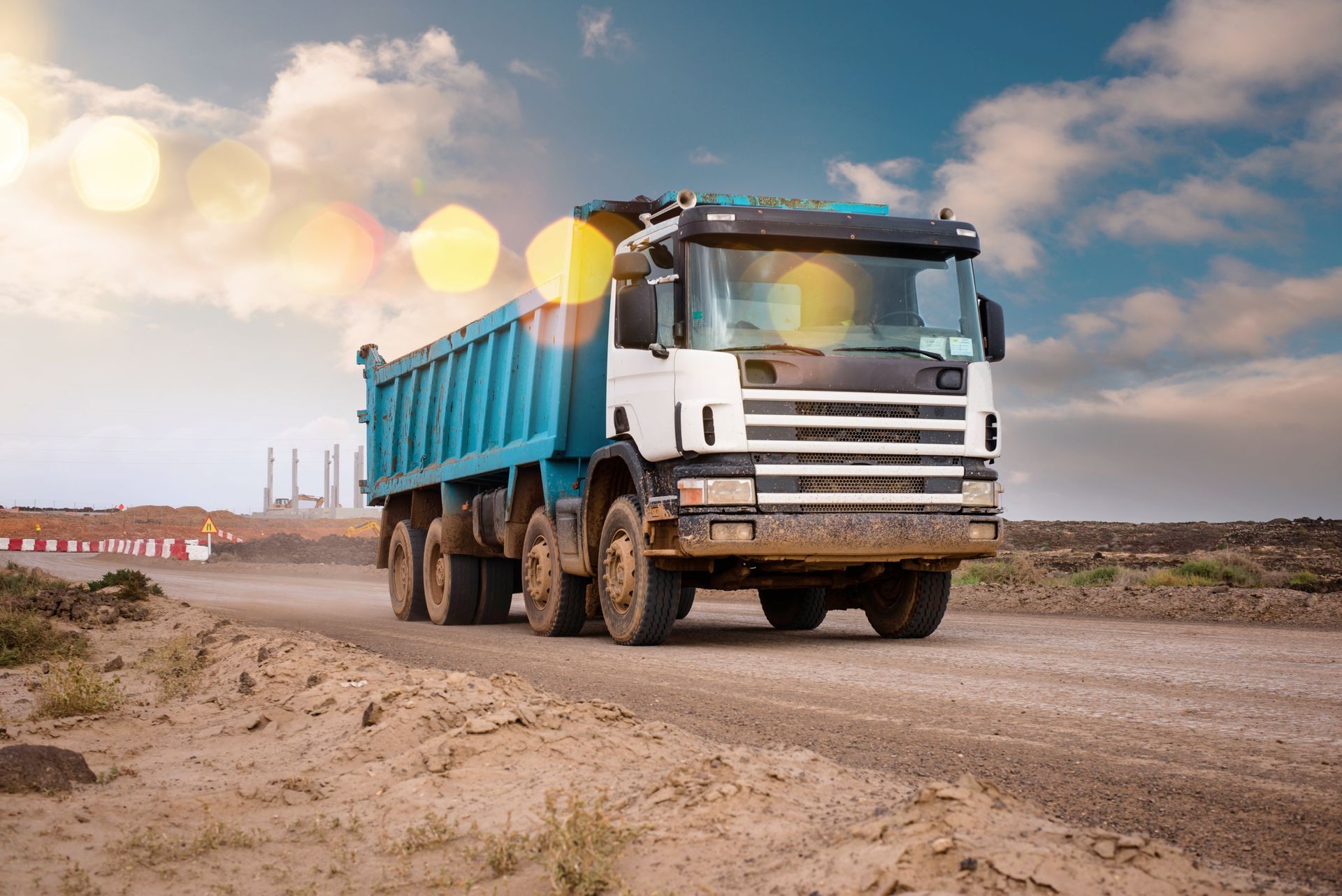 Un camion-benne blanc à benne bleue circule sur un chemin de terre sous un ciel nuageux.