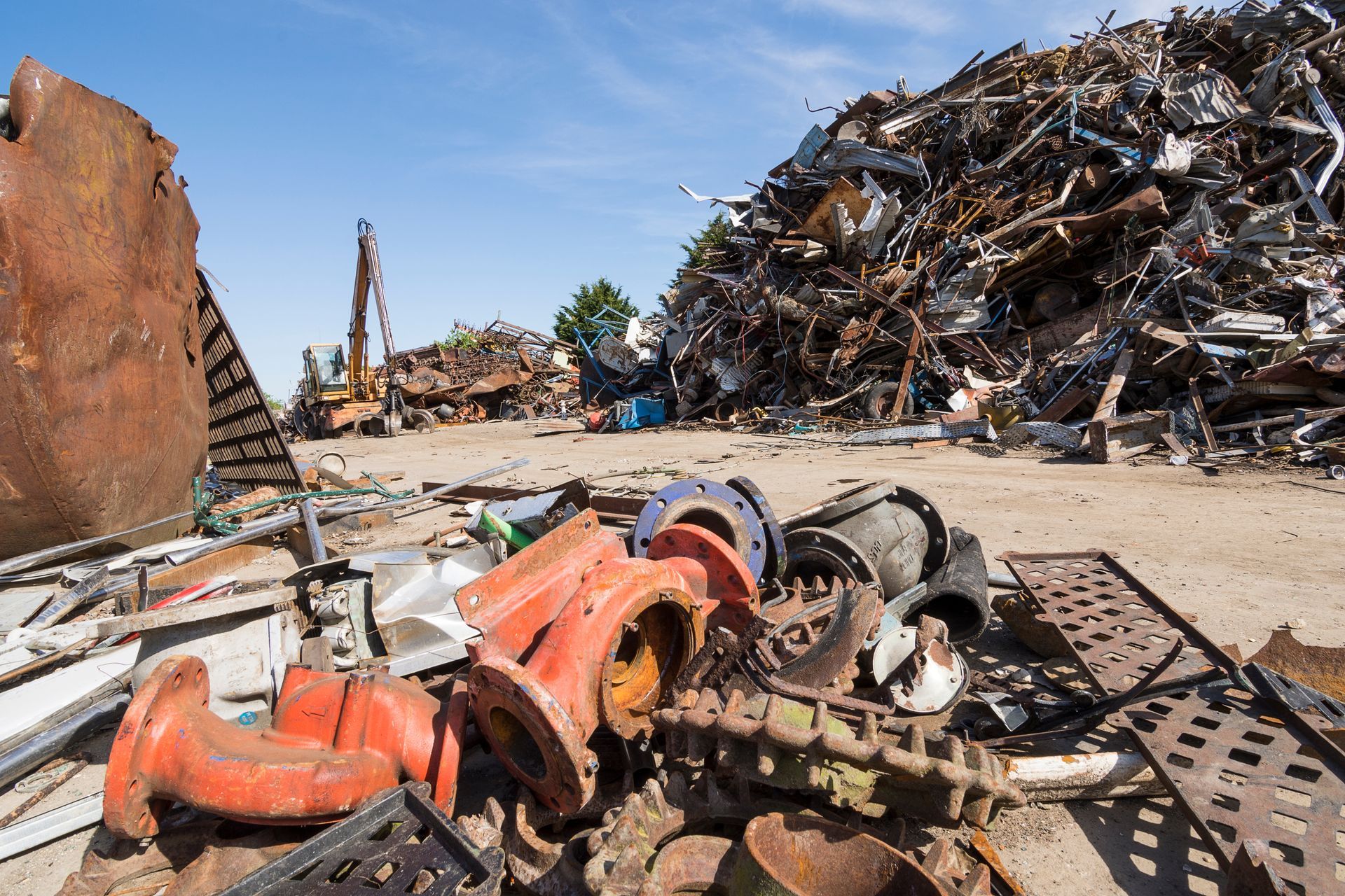 Un vaste dépôt de ferraille présente un énorme tas de pièces métalliques mises au rebut, des tuyaux industriels orange et des machines lourdes.