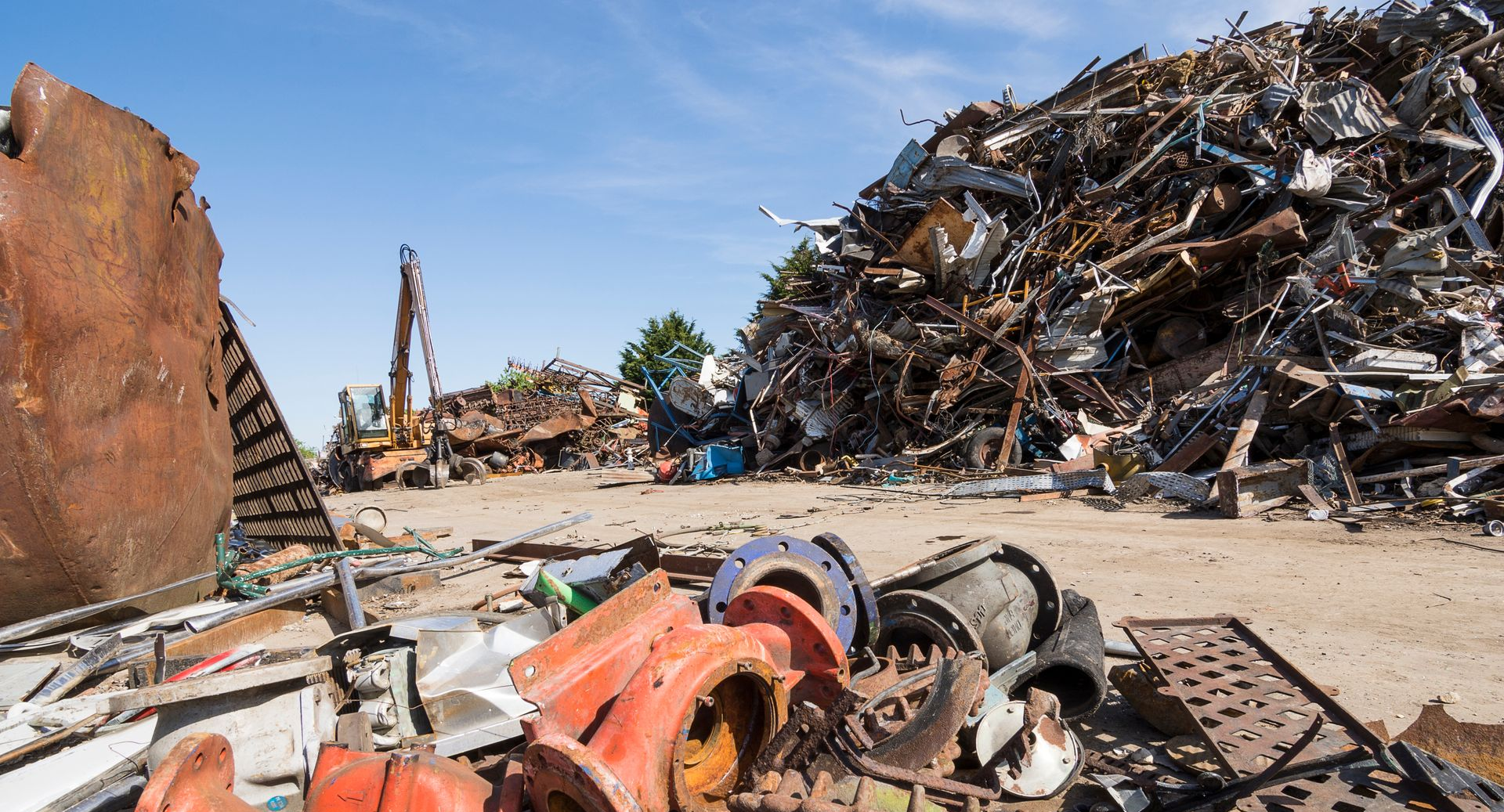 Un centre de recyclage de métaux avec de grands tas de débris, des tuyaux métalliques rouillés et une grue sous un ciel bleu azur.