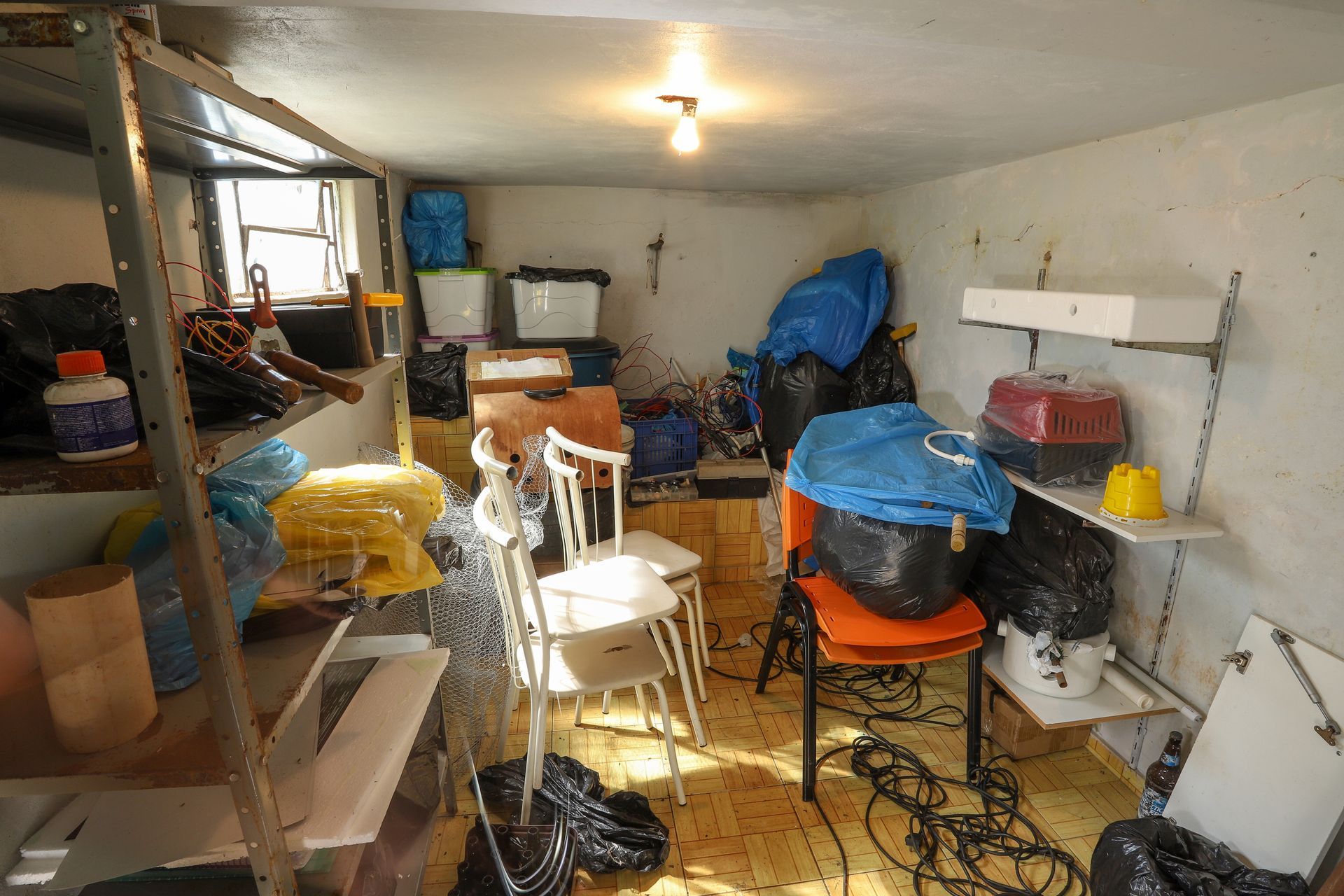 Un débarras encombré au sous-sol, avec des murs blancs, des étagères et divers conteneurs, chaises et sacs.