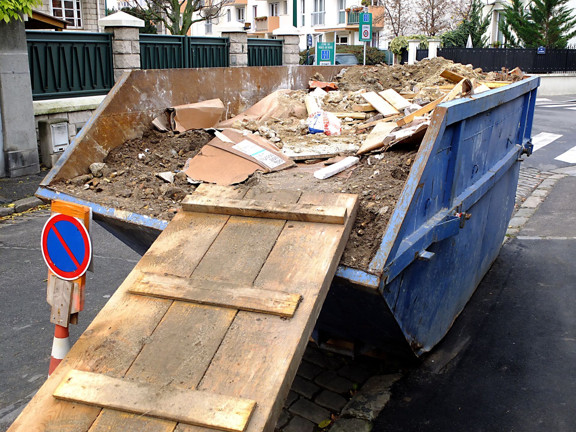 Une grande benne bleue remplie de terre et de débris de construction est garée dans une rue de la ville, accessible par une rampe en bois.
