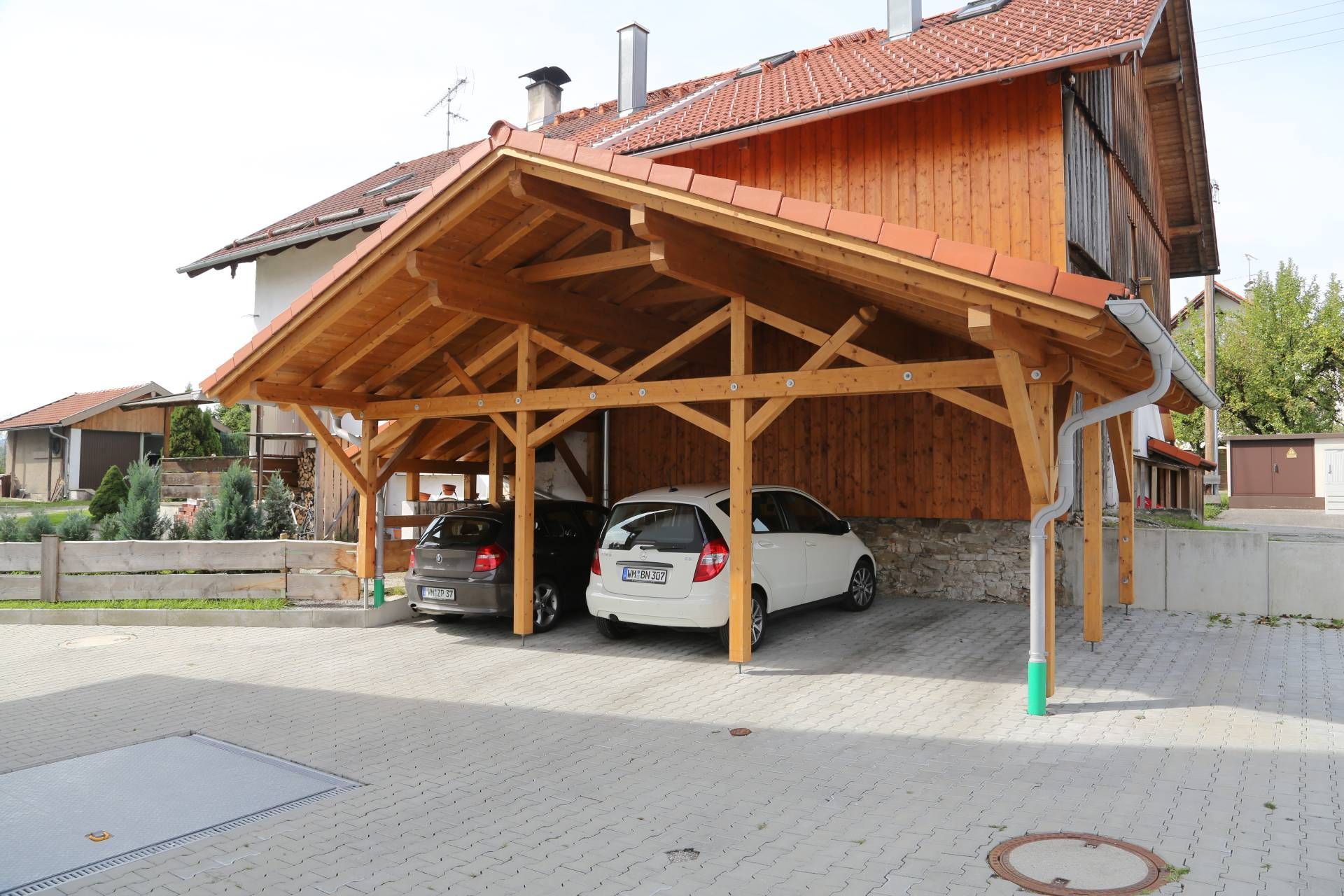 Zwei Autos parken unter einem Holzdach vor einem Haus