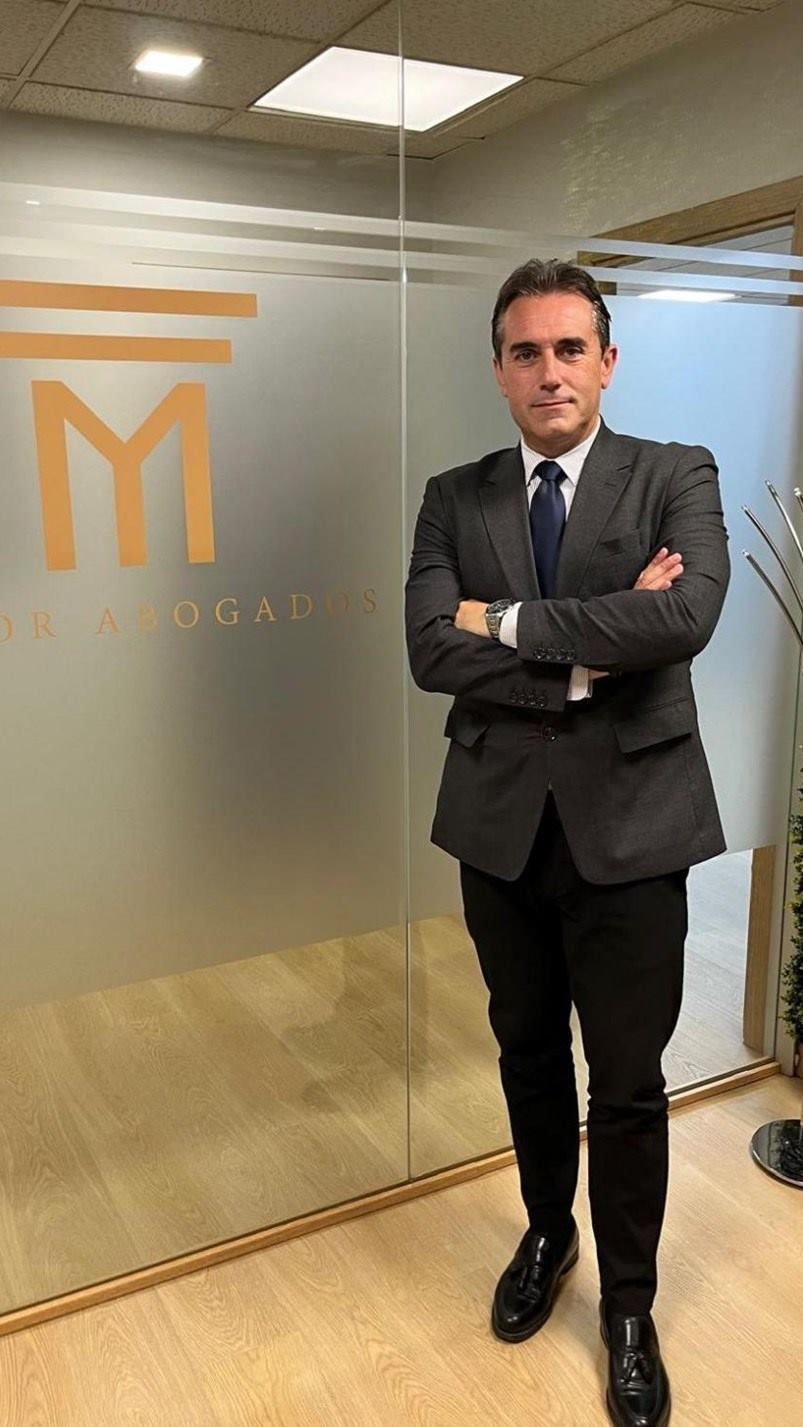 Un hombre con traje y corbata está parado frente a una pared de vidrio con los brazos cruzados.