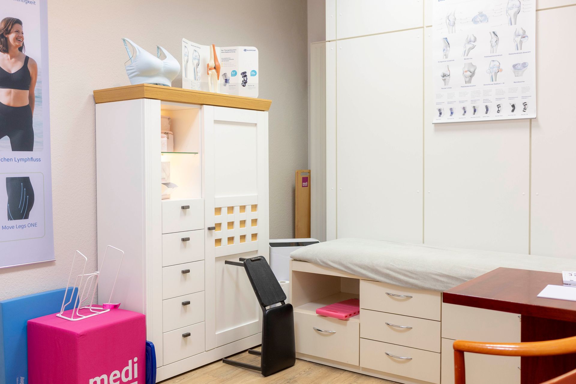 Untersuchungszimmer beim Arzt: weißer Schrank mit Vorräten, Untersuchungstisch, Poster, rosa Box und Schreibtisch.