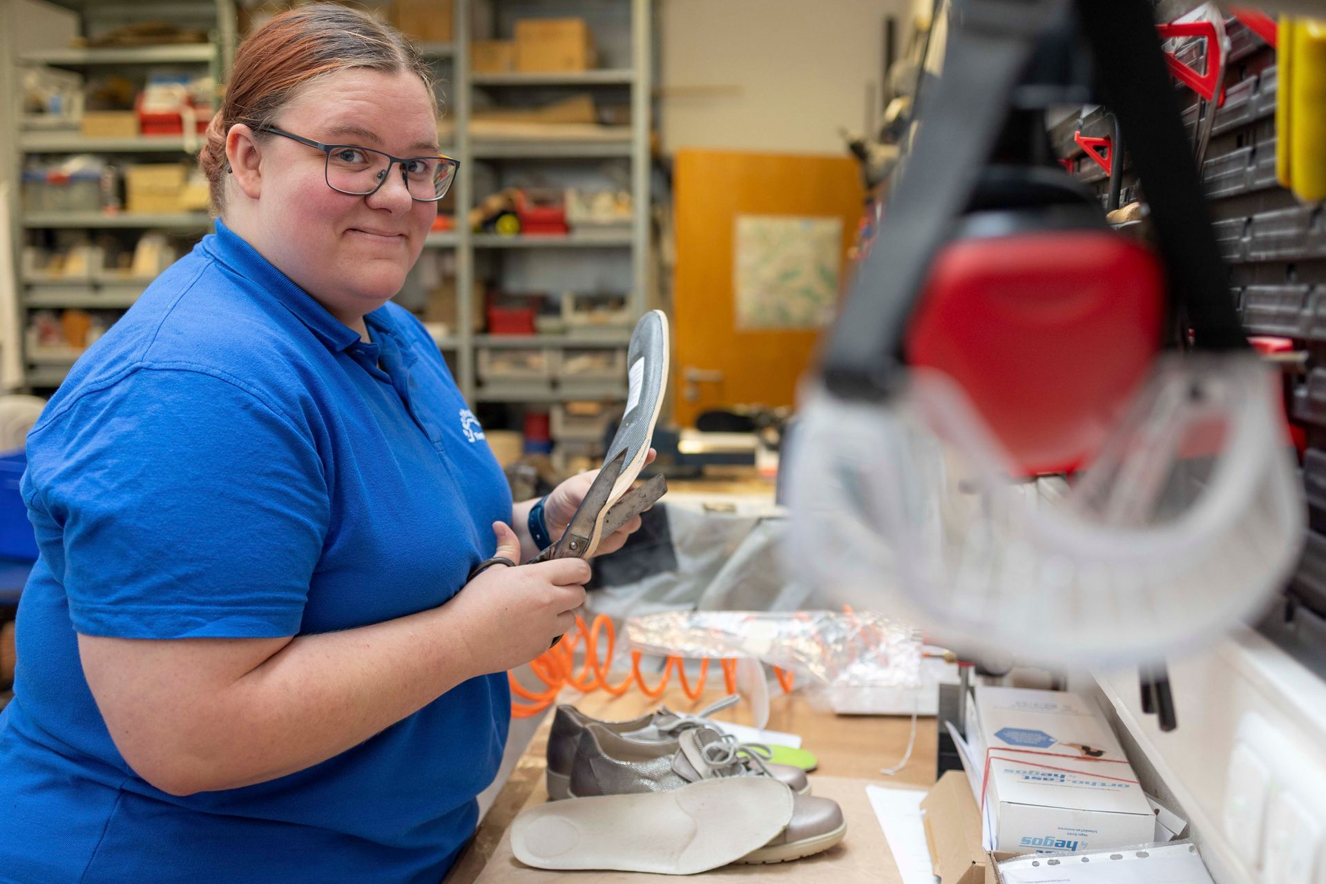 Frau im blauen Hemd lächelt, während sie in einer Werkstatt einen Schuh zusammenbaut.