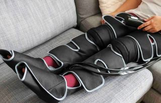 Person, die ein schwarz-graues Beinmassagegerät auf einem grauen Sofa verwendet.