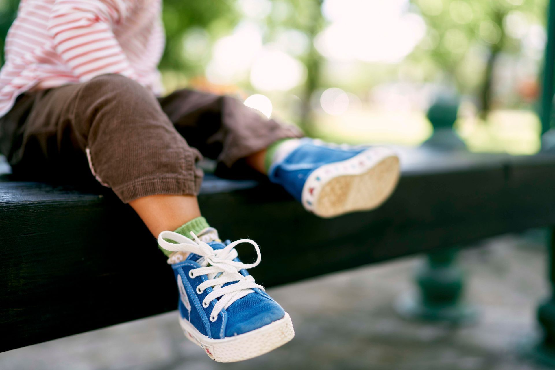 Ein Kind mit blauen Turnschuhen sitzt mit baumelnden Beinen auf einer Parkbank.