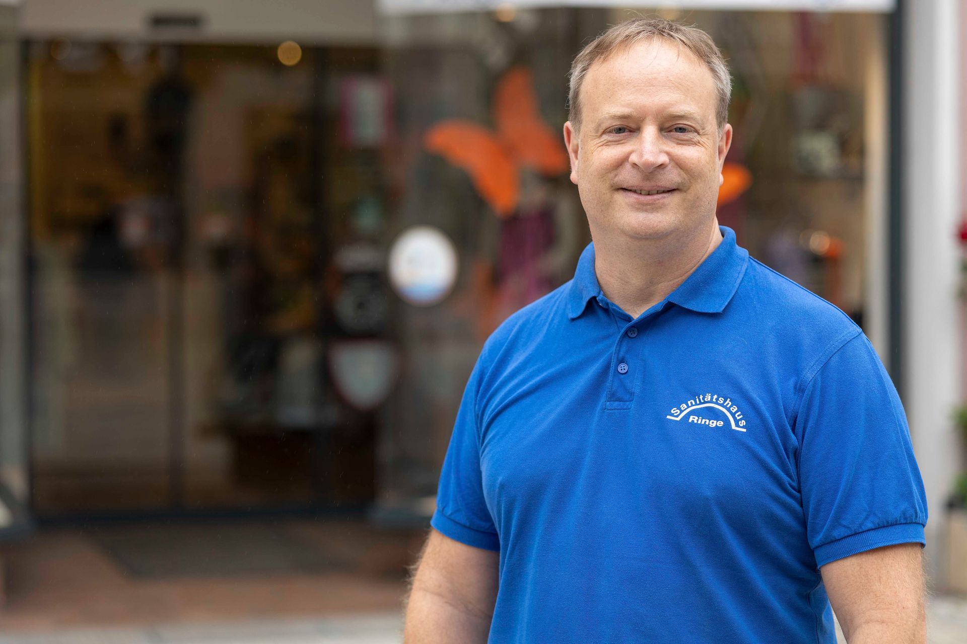 Ein Mann im blauen Poloshirt steht vor einem Geschäft mit Glastür und lächelt in die Kamera.