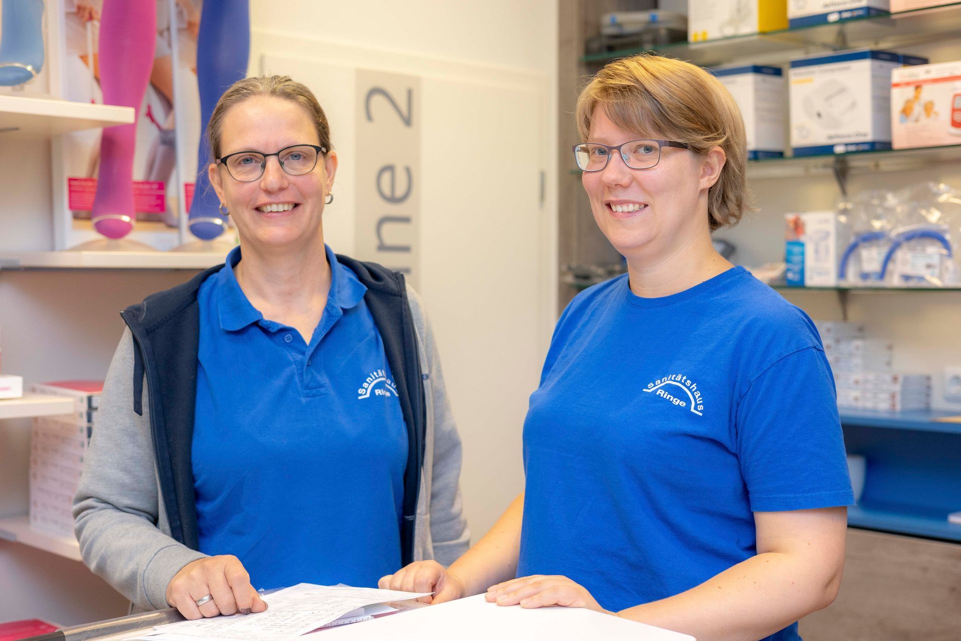 Zwei Frauen in blauen Hemden stehen lächelnd hinter einer Ladentheke.