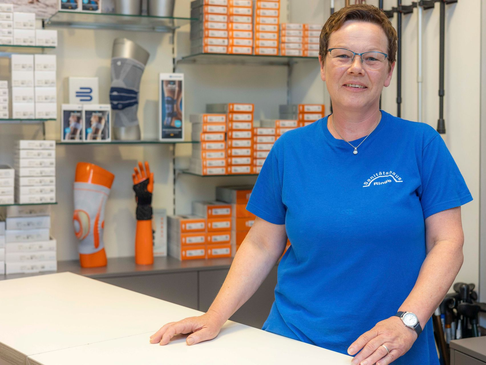 Eine Frau im blauen Hemd steht an der Theke eines Sanitätshauses, umgeben von Regalen voller Produkte.