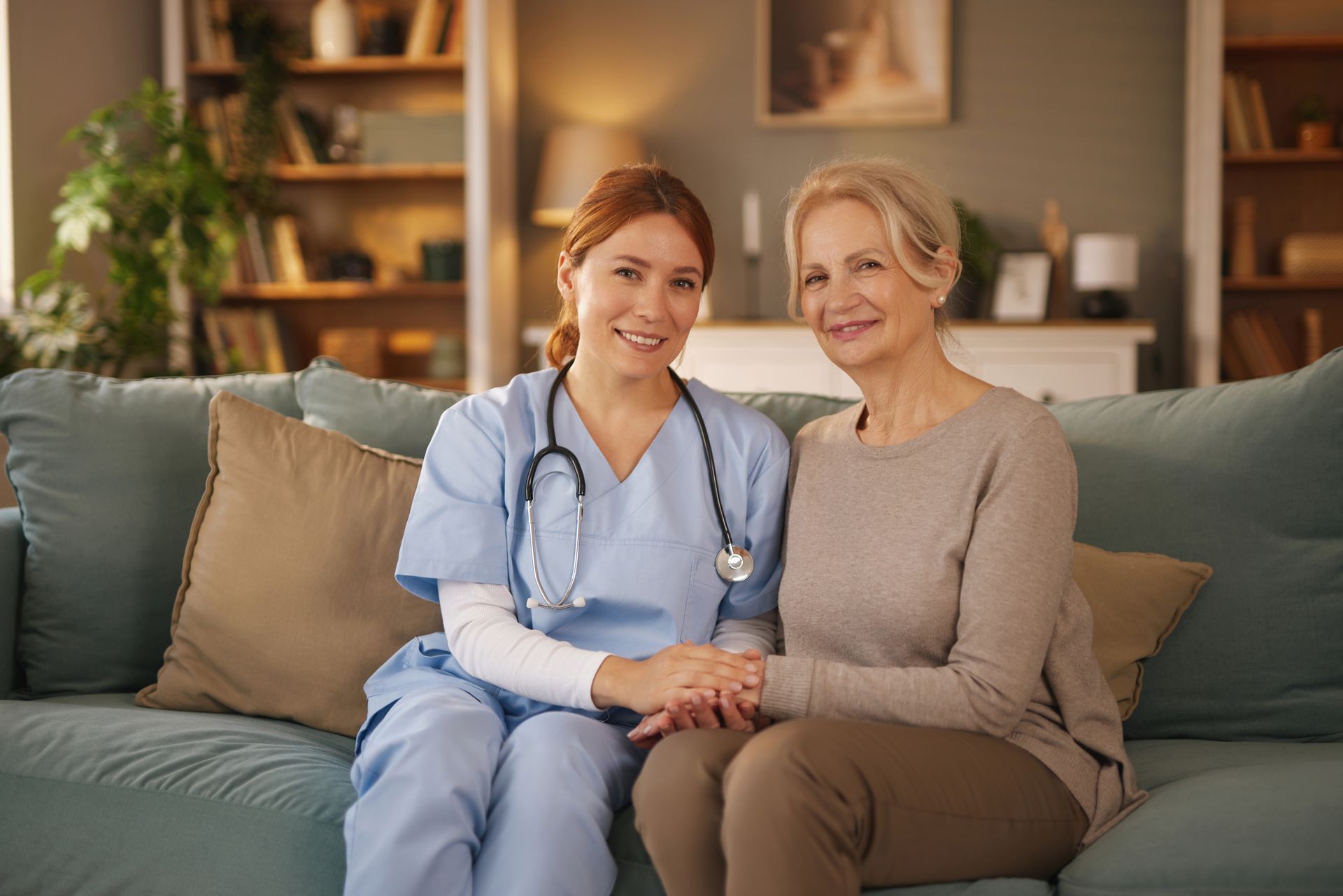 Une infirmière souriante en tenue médicale tient la main d'une femme âgée sur un canapé.