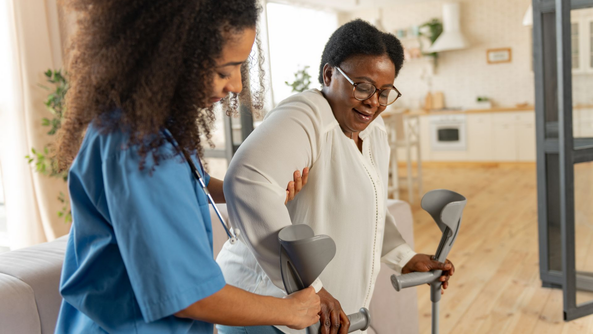 Un soignant aide une femme utilisant des béquilles à l'intérieur d'une maison ; la femme sourit.