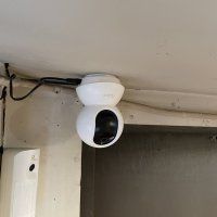 Valkoinen kattoon asennettu valvontakamera ikkunan lähellä, näkyvät johdot ja beigenvärinen seinä.