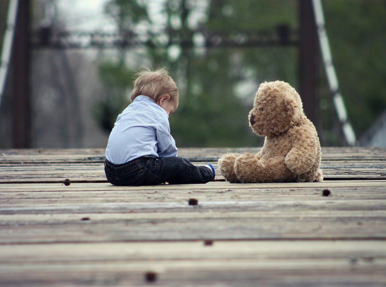 Un petit enfant et son ours en peluche sont assis face à face sur une passerelle en bois à l'extérieur.