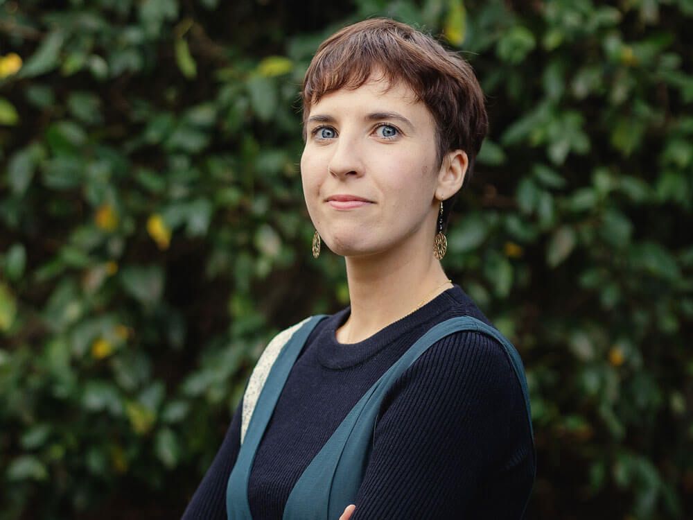 Personne aux cheveux courts, portant un pull foncé et un gilet vert, se tenant à l'extérieur devant un fond de verdure.