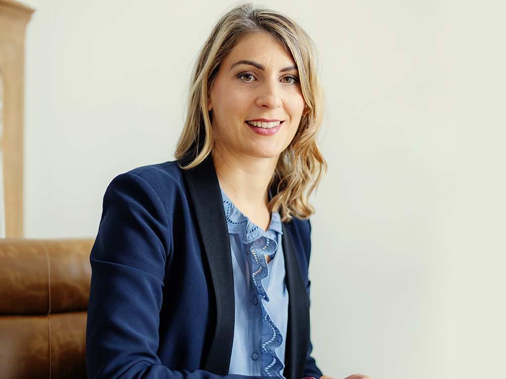 Une femme souriante, vêtue d'un blazer bleu marine, est assise sur un canapé contre un mur clair.