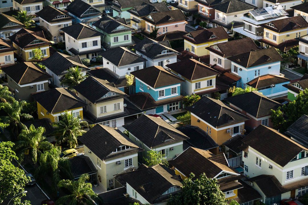 Vue aérienne de maisons de banlieue densément regroupées, aux toits sombres, et de quelques bâtiments bleu vif parmi les arbres verdoyants.