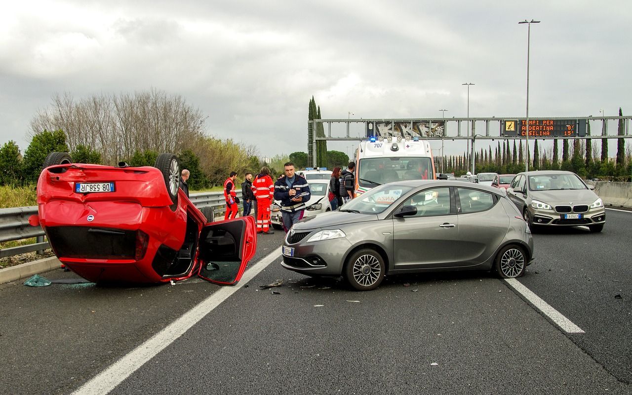 Accident impliquant plusieurs véhicules sur l'autoroute : une voiture rouge renversée, une voiture argentée au premier plan, les secours à proximité.