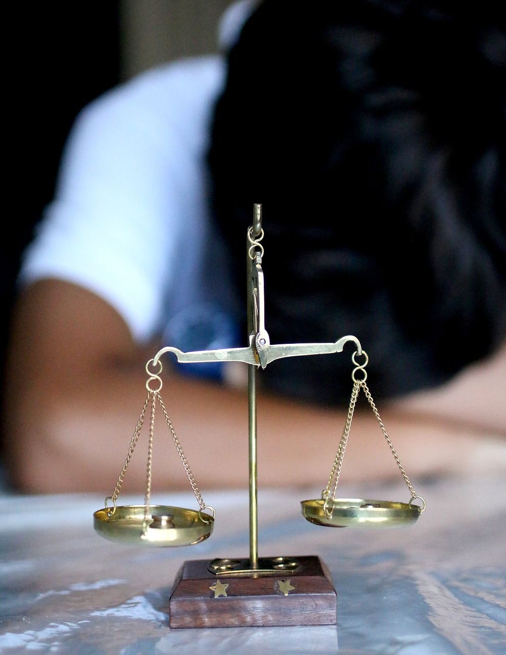 Une petite balance en laiton posée sur une table, avec une personne floue reposant sa tête en arrière-plan.