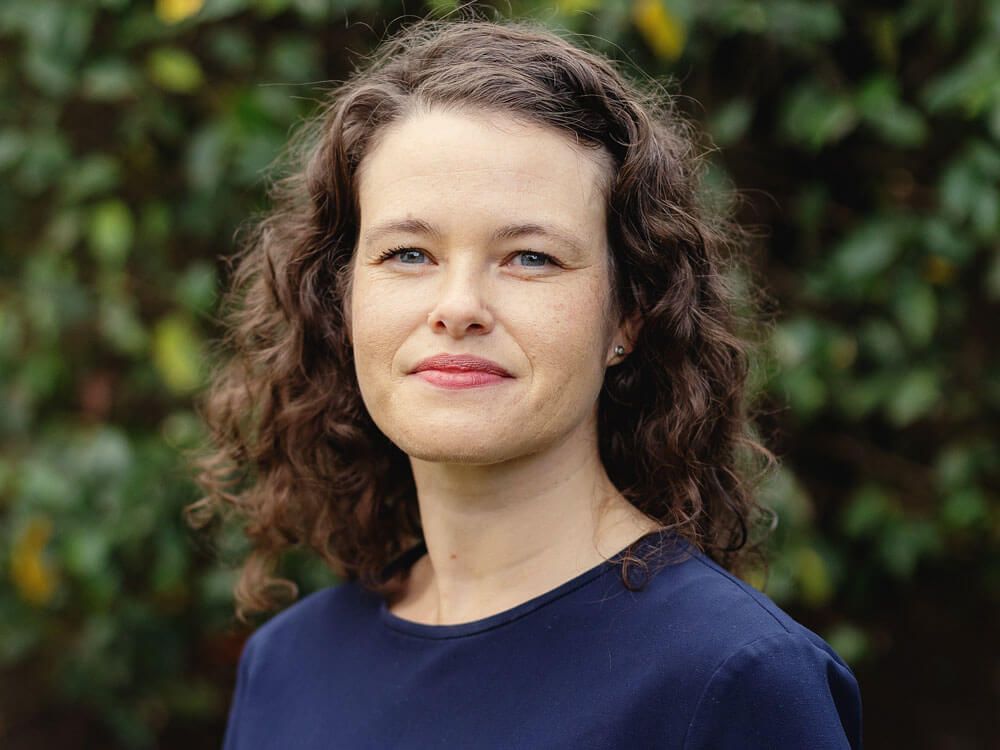 Portrait d'une personne aux cheveux bouclés portant un haut bleu marine, debout à l'extérieur devant de la verdure.
