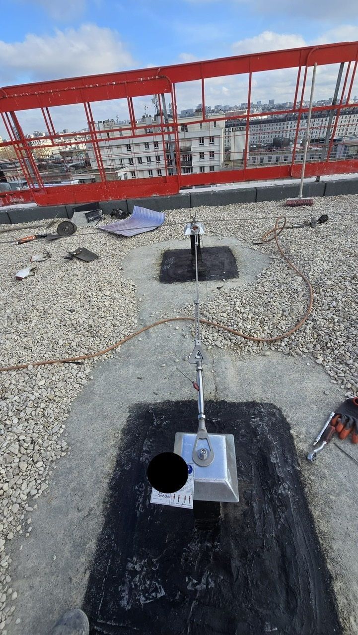 Toiture équipée d'un système de câbles de sécurité. Ancrages métalliques argentés intégrés dans un revêtement foncé. Échafaudage rouge en arrière-plan.