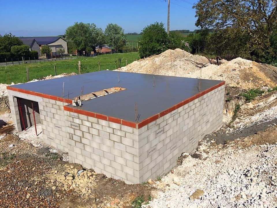 Bâtiment en parpaings partiellement construit, avec un toit plat, situé sur un terrain rural, entouré de terre et de gravier.