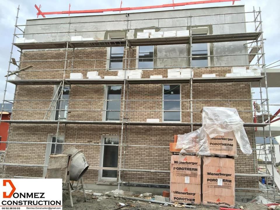 Échafaudages autour d'un bâtiment en briques en construction, avec des piles de palettes emballées devant.
