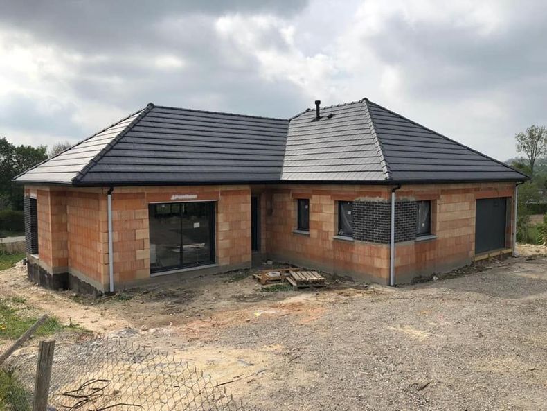 Maison de plain-pied en briques, récemment construite, avec un toit sombre, sur un terrain en terre battue sous un ciel nuageux.