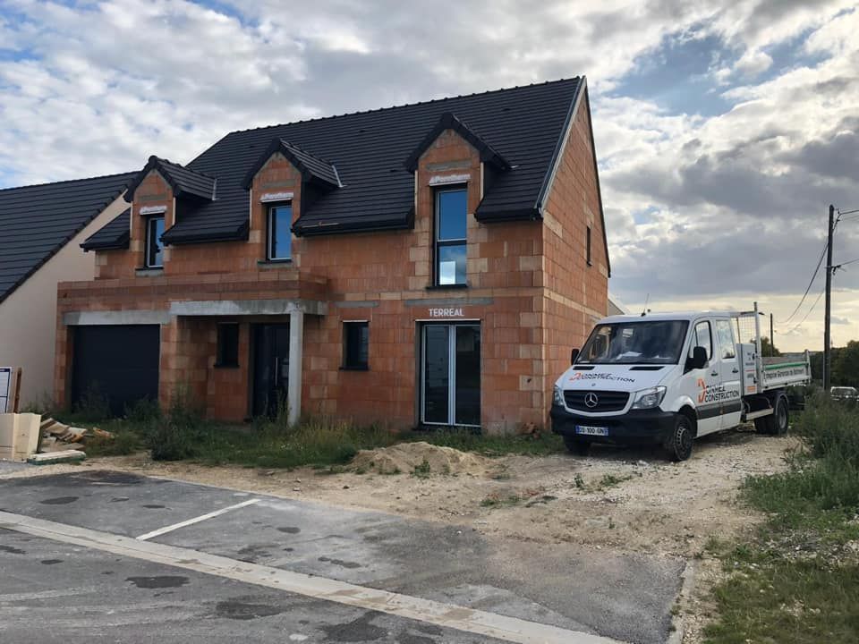 Maison en briques partiellement construite, avec une camionnette blanche garée à côté, au bord d'une route boueuse sous un ciel nuageux.