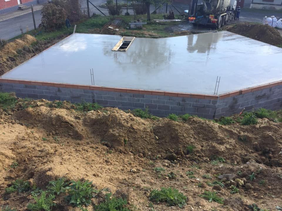 Dalle de fondation en béton frais sur un chantier, avec de la terre au premier plan et une excavatrice en arrière-plan.