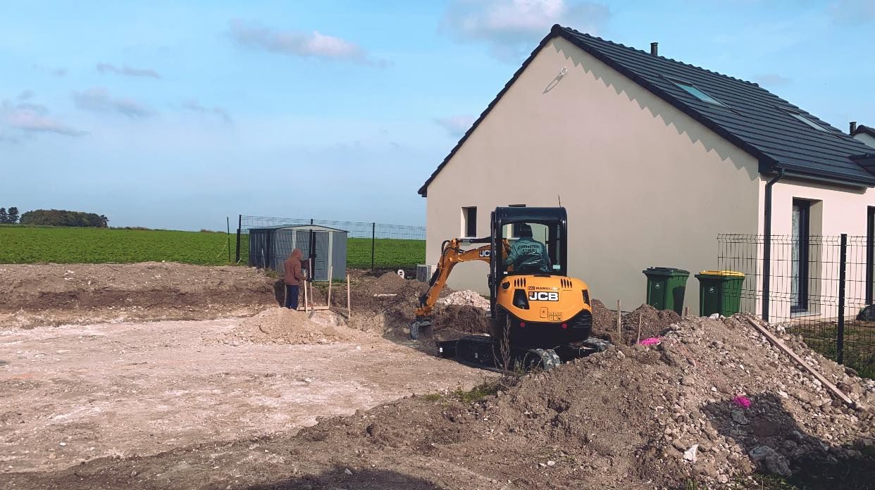 Chantier de construction avec une petite pelleteuse à côté d'une maison neuve et des tas de terre sous un ciel dégagé