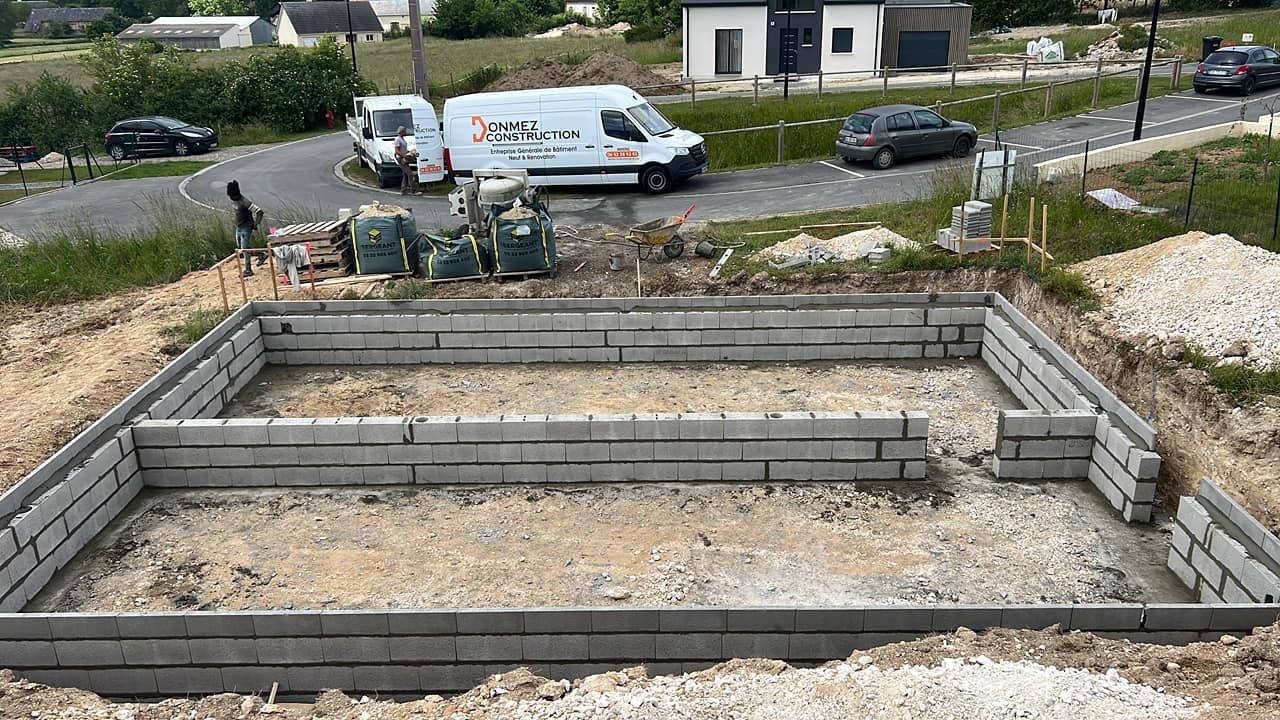 Fondation en pierre en construction, avec des ouvriers et des véhicules en arrière-plan.