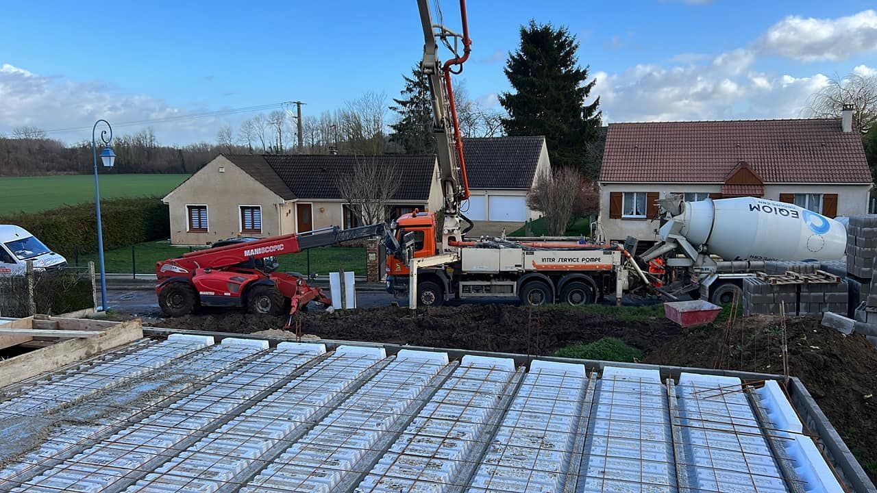 Camions et engins de chantier sur un site de construction rural, à côté d'une maison, le sol étant recouvert de neige.