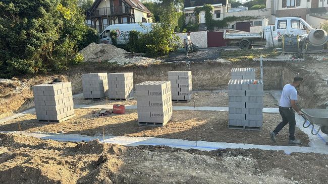 Chantier de construction avec des parpaings empilés et un ouvrier posant une bande de fondation blanche.