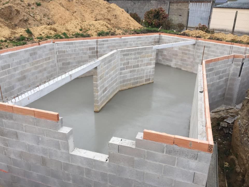Piscine ou bassin partiellement construit en parpaings, avec eaux grises et murs inachevés, sur un terrain en terre battue.