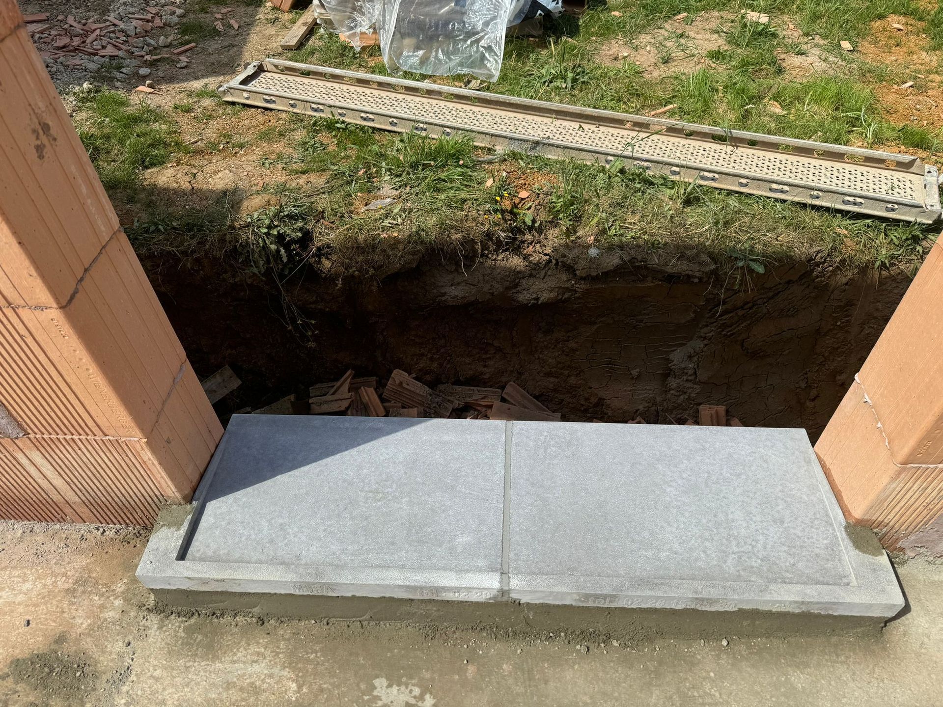 Dalle de béton enjambant une petite tranchée de terre entre deux murs de briques orange.