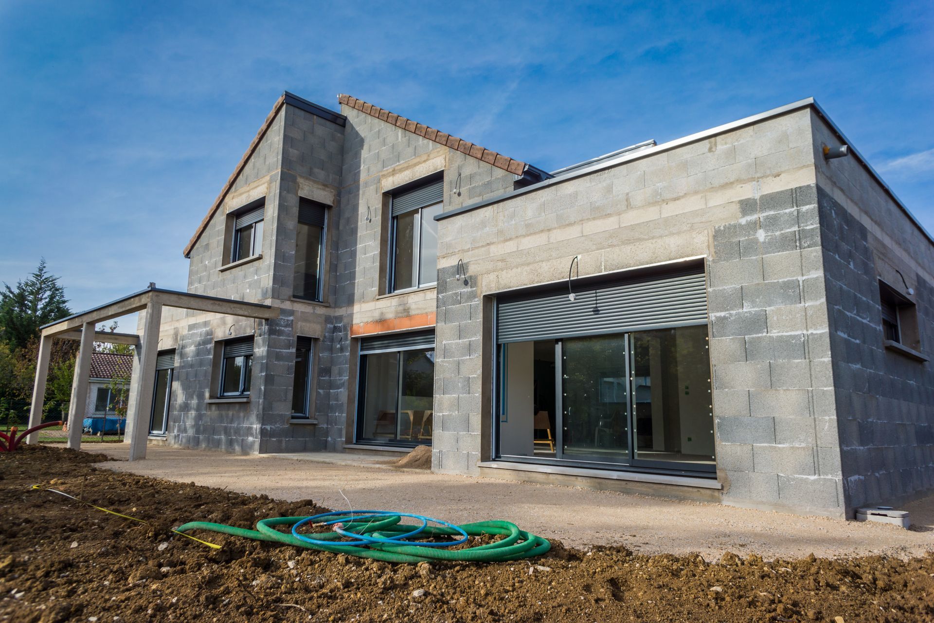 Maison en pierre grise partiellement construite, avec de grandes fenêtres et une cour inachevée sous un ciel bleu.