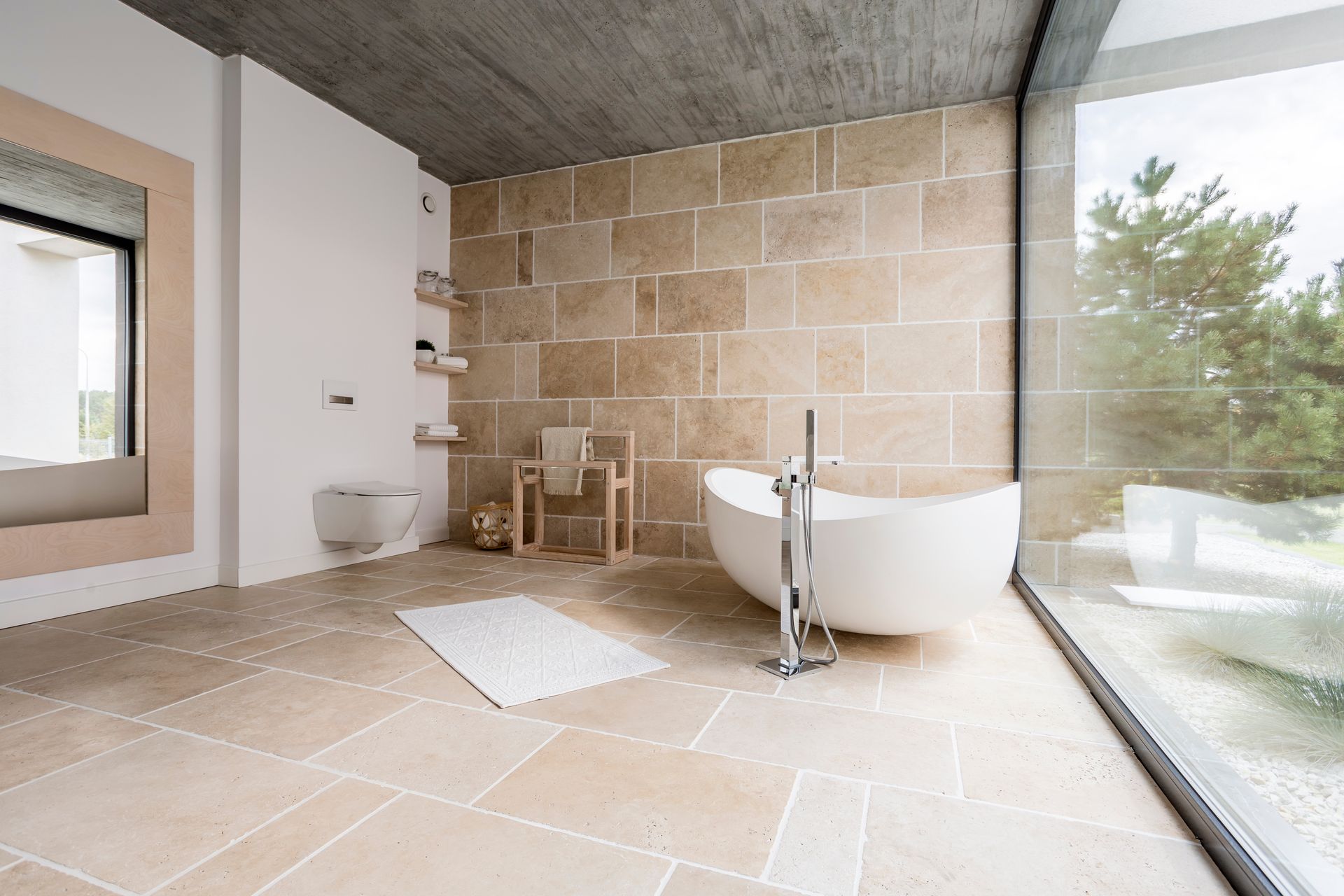 Salle de bains moderne avec baignoire îlot, murs en pierre, douche en verre et grande fenêtre donnant sur les arbres.
