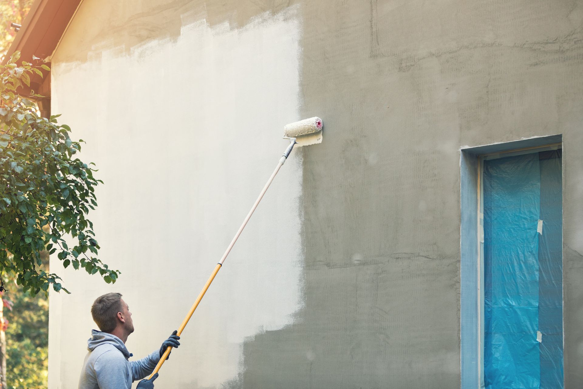 Une personne peint un mur extérieur gris au rouleau, laissant une section blanche à côté d'une porte bleue.
