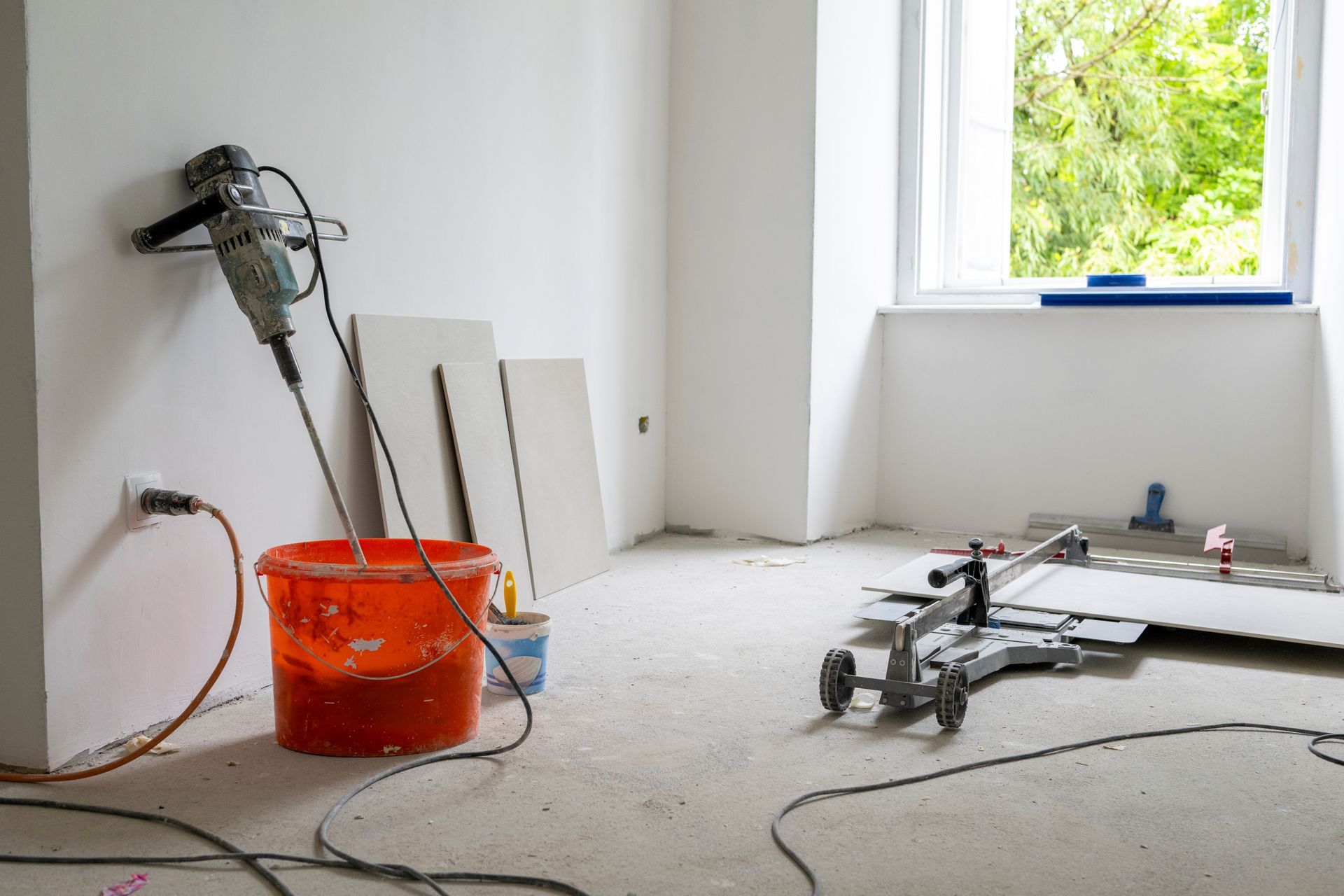 Pièce en rénovation avec outils, seau orange et panneaux de plaques de plâtre près d'une fenêtre.