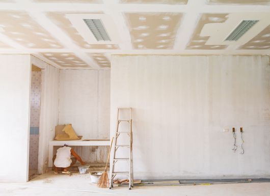 Pièce partiellement rénovée avec échelle, table, outils et plafond en plaques de plâtre non fini.