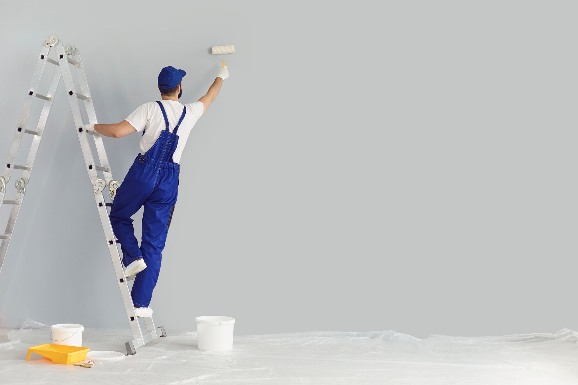 Un peintre sur une échelle peint un mur gris en blanc, avec des seaux de peinture et une bâche de protection au sol.