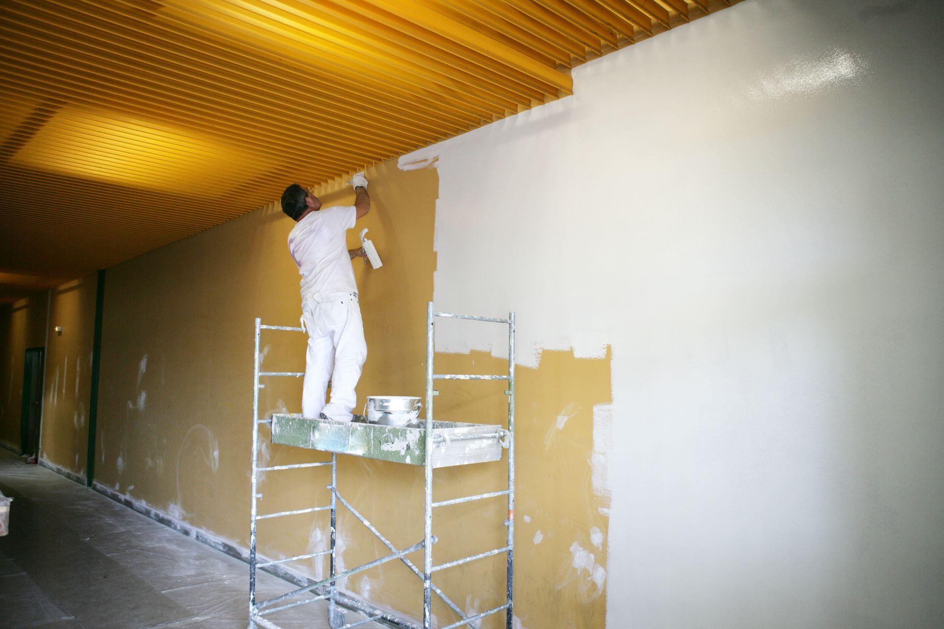 Un ouvrier peint un mur blanc depuis un échafaudage, sous un plafond jaune, dans une pièce en rénovation.