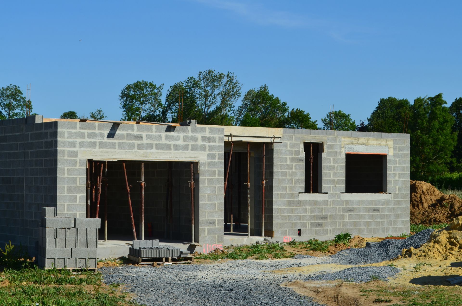 Maison en parpaings partiellement construite, avec portes et fenêtres ouvertes, sur un terrain en gravier.