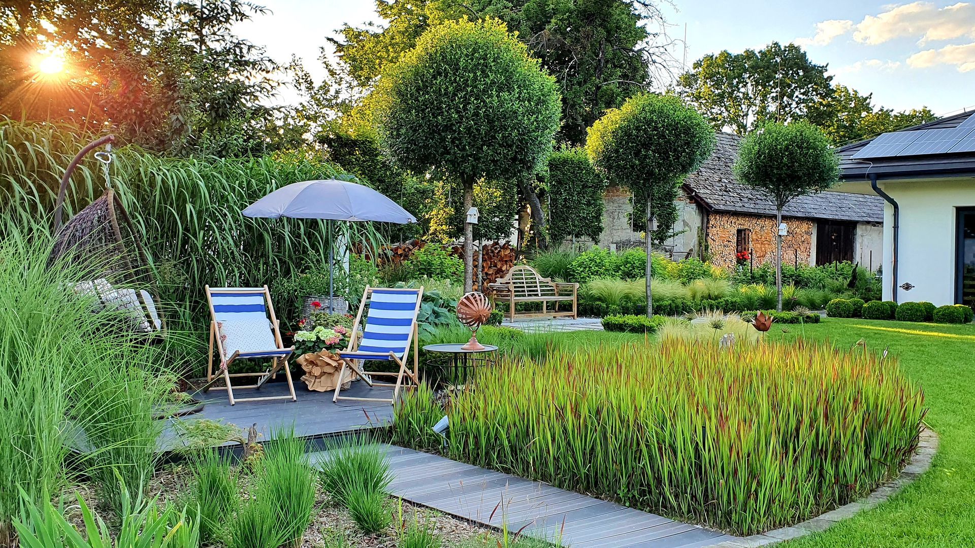 Patio de jardin luxuriant avec deux chaises bleues, un parasol blanc et un chemin de pierre au coucher du soleil.